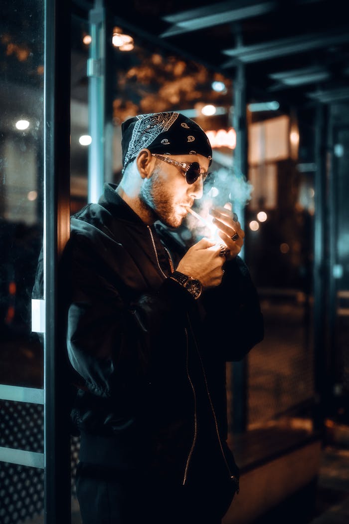 Man in a bomber jacket lights a cigarette in the city at night, exuding a moody urban vibe.