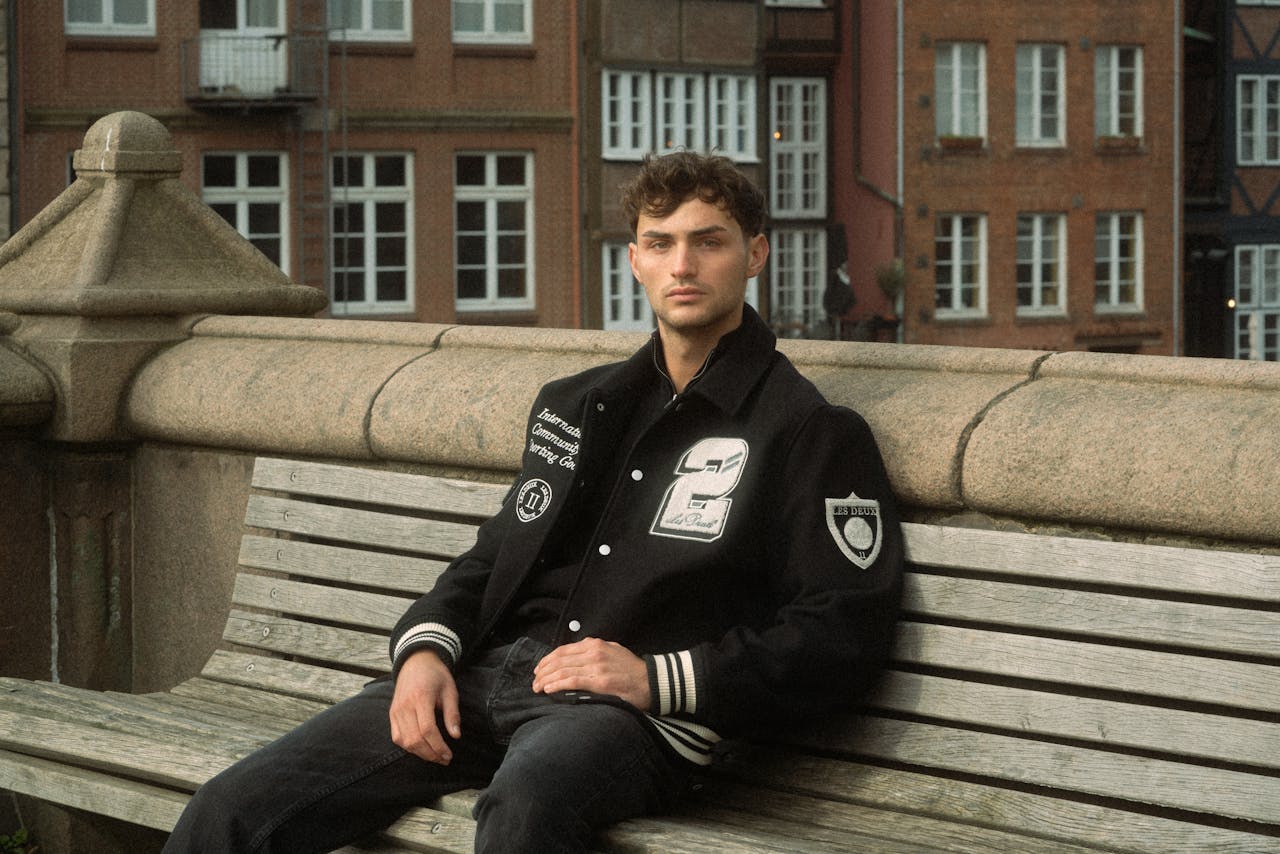 Stylish man in a varsity jacket posing on a bench with urban background.