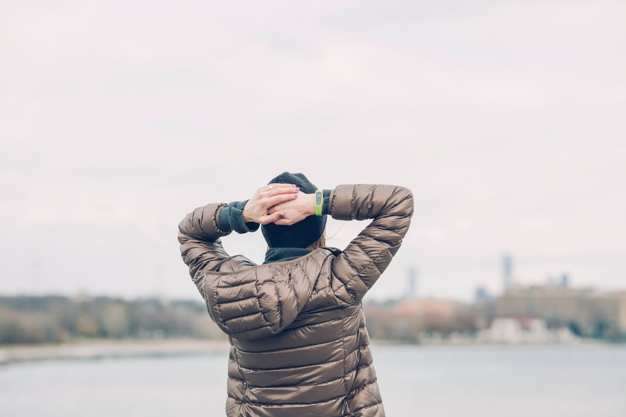 A person in a brown jacket stands with hands behind head, gazing over a blurred cityscape by the water.