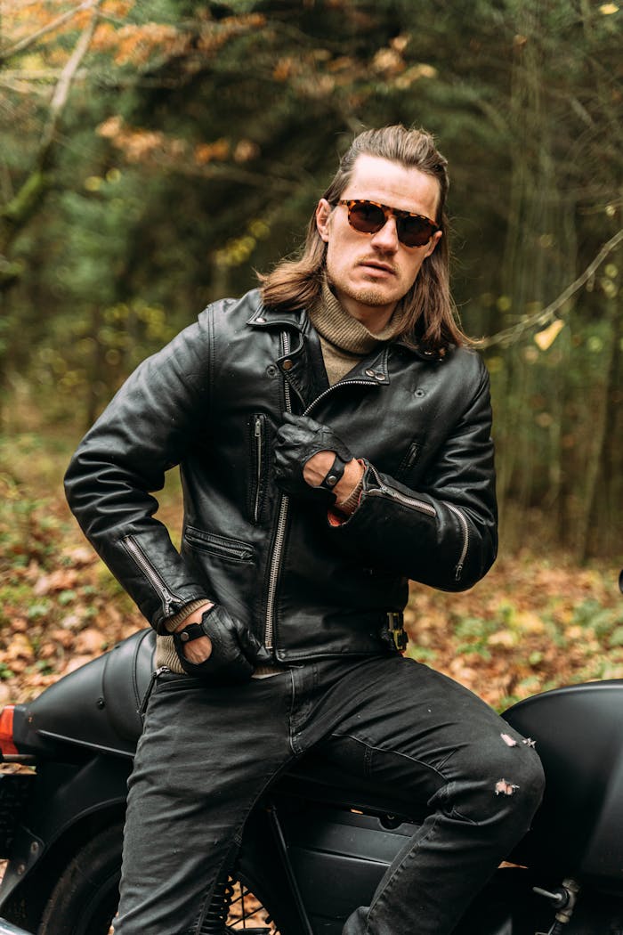 A stylish biker wearing sunglasses and leather jacket sits confidently on a motorcycle in a forest setting.