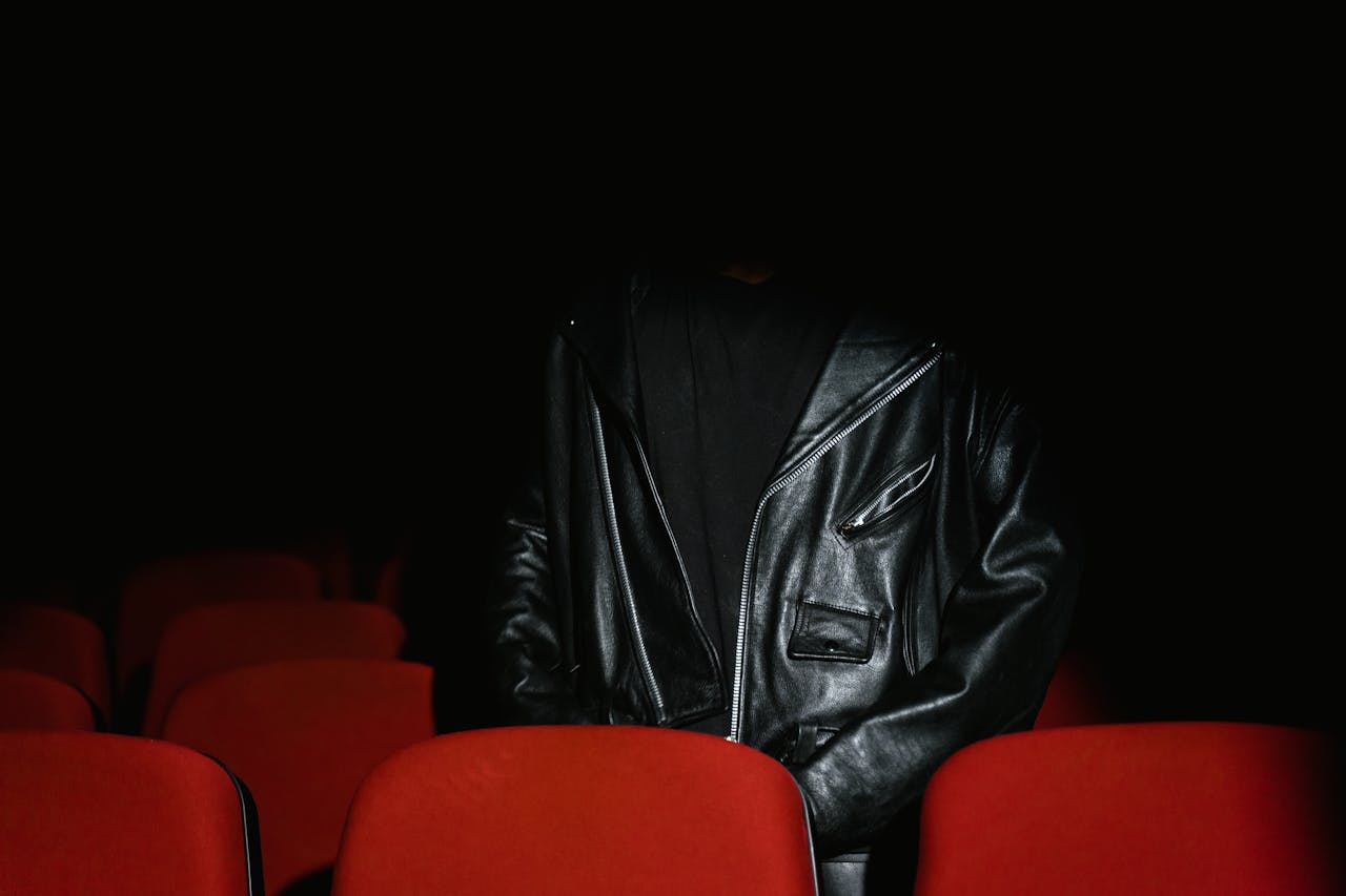 Individual wearing black leather jacket sits among red seats in a dimly lit cinema.